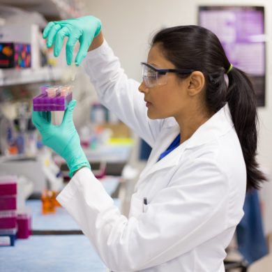 A female is seen in the lab in her white lab coat and safety glasses. She is holding a purple tray in one hand and picking up some samples with the other.