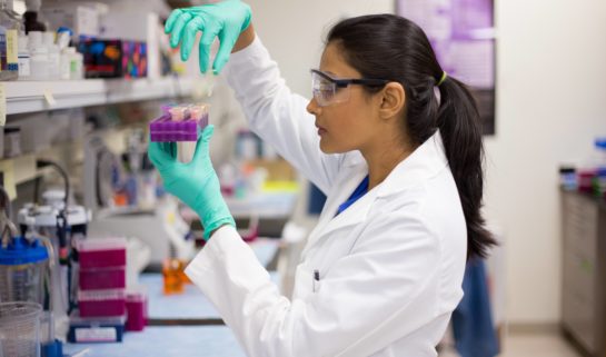 A female is seen in the lab in her white lab coat and safety glasses. She is holding a purple tray in one hand and picking up some samples with the other.