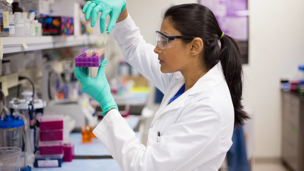 A female is seen in the lab in her white lab coat and safety glasses. She is holding a purple tray in one hand and picking up some samples with the other.