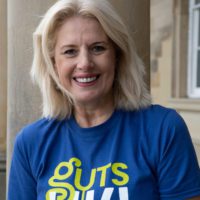 Helen has short blonde hair and is wearing a blue t-shirt that says Guts UK in green and white. She is outside and a building with windows is visible behind her.