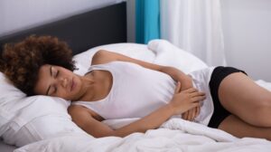 A woman is lying on top of a bed. She looks sad, with her hands clasped over her stomach, indicating pain or discomfort.