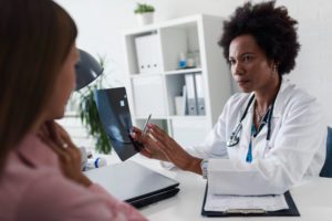 A woman wearing a white lab coat is holding a scan and showing it to a woman wearing a pink shirt. They are both sat down in a room with a clipboard of papers in between them.
