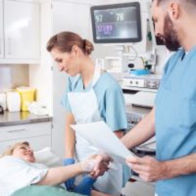 A lady is laid on the bed with a smile in the hospital. There are two male and female healthcare professionals standing and facing her. She has her right hand reaching out to shake a male healthcare professional who is wearing light blue scrubs. The female healthcare professional is wearing an apron over her light blue uniform.