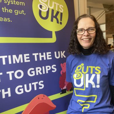 Melanie has short, brown curly hair and dark round glasses. She is wearing a blue shirt that says Guts UK in green and white. She is stood next to a blue pop up banner that shows the Guts UK logo and says 'It's time the UK got to grips with guts.'