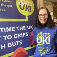 Melanie has short, brown curly hair and dark round glasses. She is wearing a blue shirt that says Guts UK in green and white. She is stood next to a blue pop up banner that shows the Guts UK logo and says 'It's time the UK got to grips with guts.'