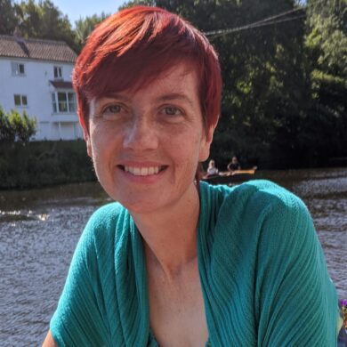 Fiona has red hair and wears a green top. She is sat outdoors and is smiling at the camera.
