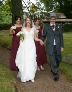 Derek holding Donna's hand on her wedding day. Donna is a in a white bridal dress. Behind them is 2 bridesmaids wearing red dresses