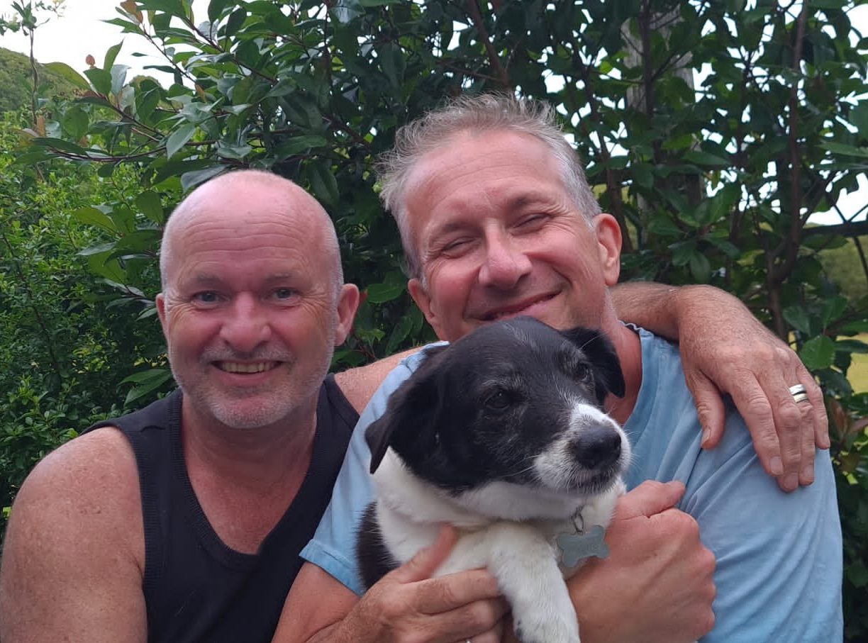 Teym has his arm around Scott, they are smiling. Scott is holding a dog.