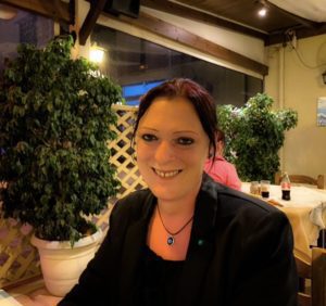 Sue in a black top, black blazer, with a necklace around her neck. Her hair is tied back, smiling for the camera. Sat in a restaurant.