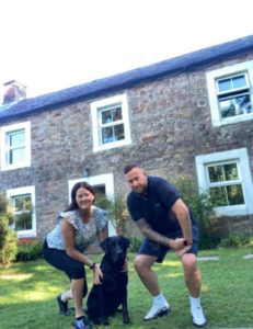 Jamie and his partner Donna with their black Labrador, Kai, in front of their house. 