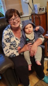 Anne wearing a white and blue top with one of her grandchildren sat on her lap. Smiling at the camera.