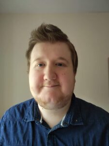 A selfie of Michael smiling gently in front of a plain light wall. He has short brown hair styled upwards, light eyes and is wearing a blue denim shirt.