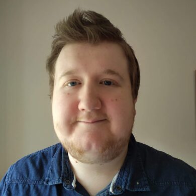 A selfie of Michael smiling gently in front of a plain light wall. He has short brown hair styled upwards, light eyes and is wearing a blue denim shirt.
