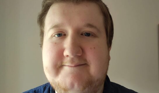 A selfie of Michael smiling gently in front of a plain light wall. He has short brown hair styled upwards, light eyes and is wearing a blue denim shirt.