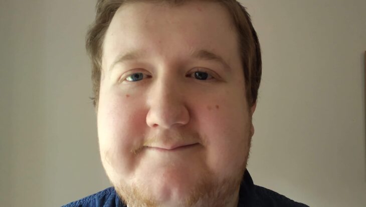 A selfie of Michael smiling gently in front of a plain light wall. He has short brown hair styled upwards, light eyes and is wearing a blue denim shirt.