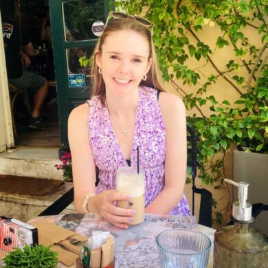 Ellie is sat outside, at a tabvle. She has a drink in her hand and is smiling at the camera. She is wearing a pink vest top and has sunglasses on top of her head.
