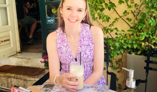 Ellie is sat outside, at a tabvle. She has a drink in her hand and is smiling at the camera. She is wearing a pink vest top and has sunglasses on top of her head.