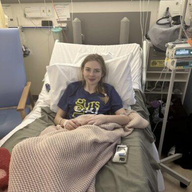 Elle is smiling and sat up on a hospital bed wearing a Guts UK t-shirt.