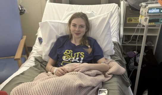 Elle is smiling and sat up on a hospital bed wearing a Guts UK t-shirt.