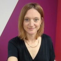 Victoria is standing in front of a pink and white wall. She is wearing a black top and a gold necklace and has shoulder length hair.