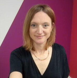 Victoria is standing in front of a pink and white wall. She is wearing a black top and a gold necklace and has shoulder length hair.