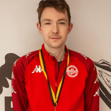 Paul wears a red jacket and is smiling at the camera. He also has a medal on a yellow and black lanyard around his neck.