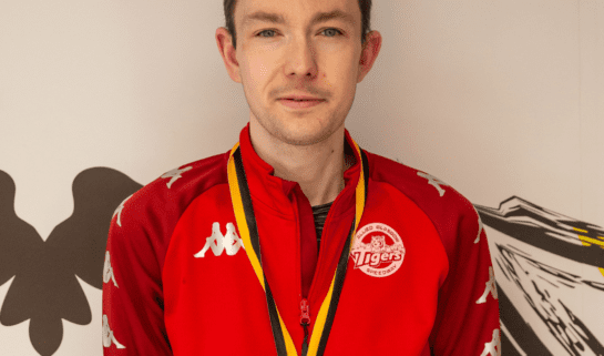 Paul wears a red jacket and is smiling at the camera. He also has a medal on a yellow and black lanyard around his neck.