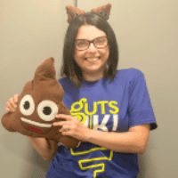 Cherylyn has short dark hair and black glasses. She is wearing a blue t-shirt that says Guts UK in green and white. She is wearing poo boppers on her head and is holding a poo cushion with eyes and a smiley face.