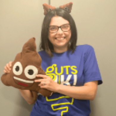 Cherylyn has short dark hair and black glasses. She is wearing a blue t-shirt that says Guts UK in green and white. She is wearing poo boppers on her head and is holding a poo cushion with eyes and a smiley face.