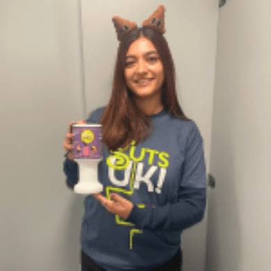 Leela has long brown hair. She is wearing a blue Guts UK t-shirt with the charity's green and white logo on it. She has a long sleeved blue top underneath. She is also wearing poo boppers on her head. She is holding a collection tin in her hands.