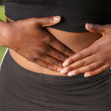 A person wearing a black crop top and black leggings shows their stomach and has both of their hands placed on either side of it, with one pressing into the stomach indicating pain or discomfort.