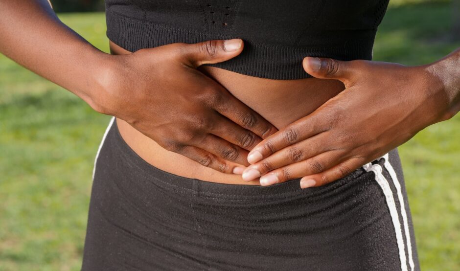 A person wearing a black crop top and black leggings shows their stomach and has both of their hands placed on either side of it, with one pressing into the stomach indicating pain or discomfort.