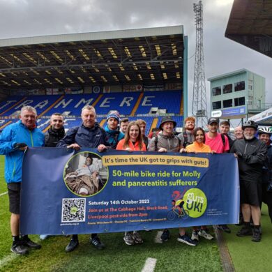 A group of people are holding the Guts UK banner while smiling at the camera. They are at a football stadium.