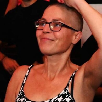 Cheryle is at a rave, dancing, with one arm up in the air. She is smiling and wears a dogtooth print vest top and black framed glasses. The photo is cropped from her waist upwards.