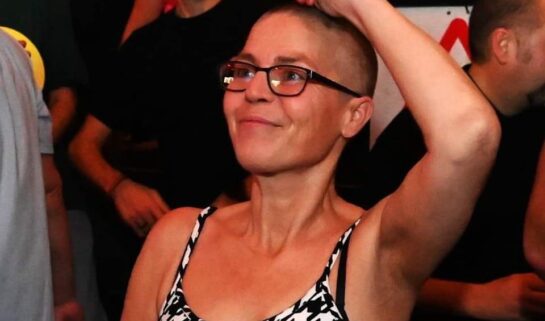 Cheryle is at a rave, dancing, with one arm up in the air. She is smiling and wears a dogtooth print vest top and black framed glasses. The photo is cropped from her waist upwards.
