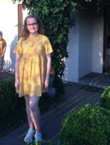 Dominika is wearing a yellow dress with flowers on and is stood, smiling at the camera. She is wearing white. lace-up trainers and is standing in front of green foliage. 