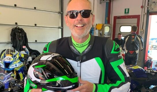 Mark is standing in a garage. He is wearing black, green and white motorbike leathers and is holding a matching helmet. He is smiling at the camera and wears black sunglasses. There is a motorbike behind him.