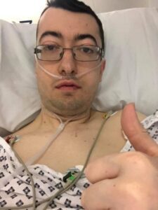 Matt is in the hospital and taking a selfie while he is laid on the hospital bed. He is wearing a hospital gown and has multiple tubes attached to him while holding a thumbs up. He stares at the camera without any emotions on his face.