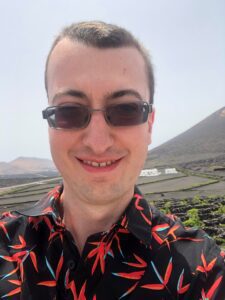Matt is taking a selfie with a big smile on his face. He is wearing black sunglasses and a black shirt with multiple orange dragonflies on it. Green fields are seen behind him.