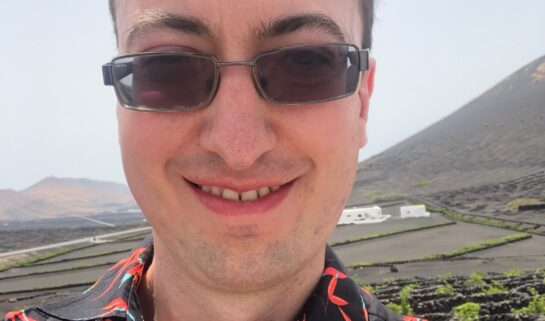 Matt is taking a selfie and has a big smile on his face. He is wearing transition glasses and a black shirt with multiple orange dragonflies on it. Green fields are seen behind him.