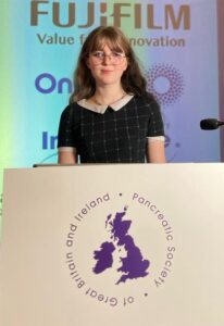 Lola stands at the speech podium ready to talk to a room full of healthcare professionals. She is looking at the camera and smiling.