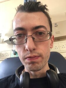 Matt is taking a selfie of his face while he is sat on a navy chair in the hospital. He has black headphones around his neck and is staring emotionless at the camera. 