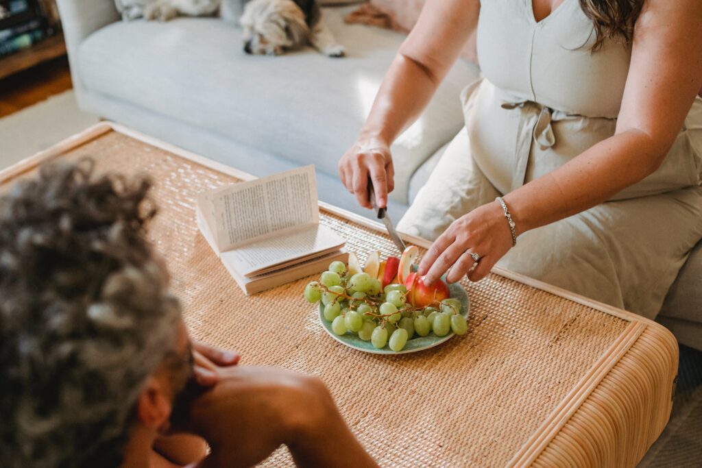 Woman cutting a plate of fruit up, sat on a sofa. The plate is rested on the table in front of her.