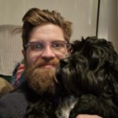 James is smiling at the camera with his dog to the right. The photo is cropped to show the top of his arms upwards. He is wearing a black jumper, has ginger hair and a beard. He is also wearing glasses. His dog is black and curly, with a white patch on its chest and a few white whiskers.
