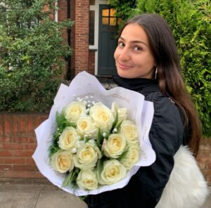 Alexandra is smiling towards the camera. The photo is taken from a side view and her body is facing left. She has a bunch of white roses in her arms which she is proudly showing the camera, with a house in the background. 