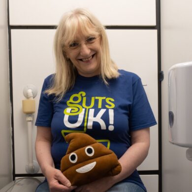 Julie is sat on the toilet wearing dark blue denim jeans (that are pulled up!) and a Guts UK t-shirt. She is holding a poo emoji cushion and is smiling at the camera.