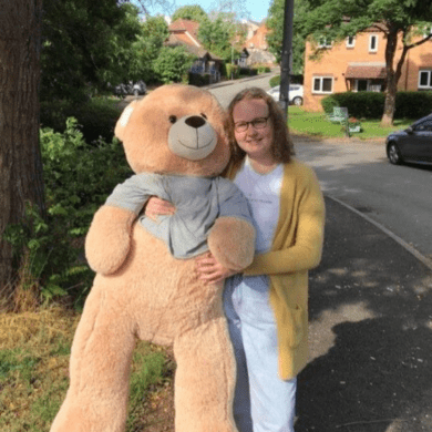 Dominika is wearing a white t-shirt, light denim jeans and a yellow cardigan. She also is wearing black rimmed glasses and is smiling at the camera, holding a light brown giant teddy bear.