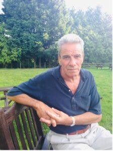 John in 2013, aged 61, sat on a park bench wearing a blue navy shirt looking at the camera.