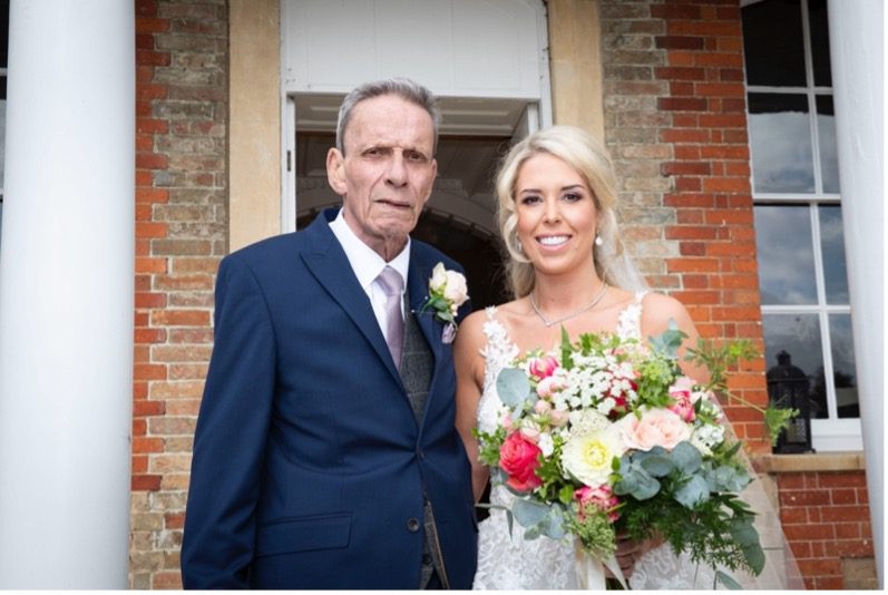 John is stood beside Charlotte in his navy suit. Charlotte is wearing a white laced wedding dress and holding a bouquet of flowers. Charlotte is smiling at the camera.