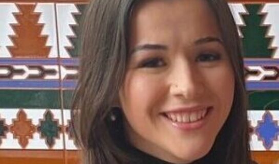 Sophie is looking at the camera smiling and has her hair down. She is wearing a black turtleneck long-sleeved top sat in front of a patterned tiled wall.